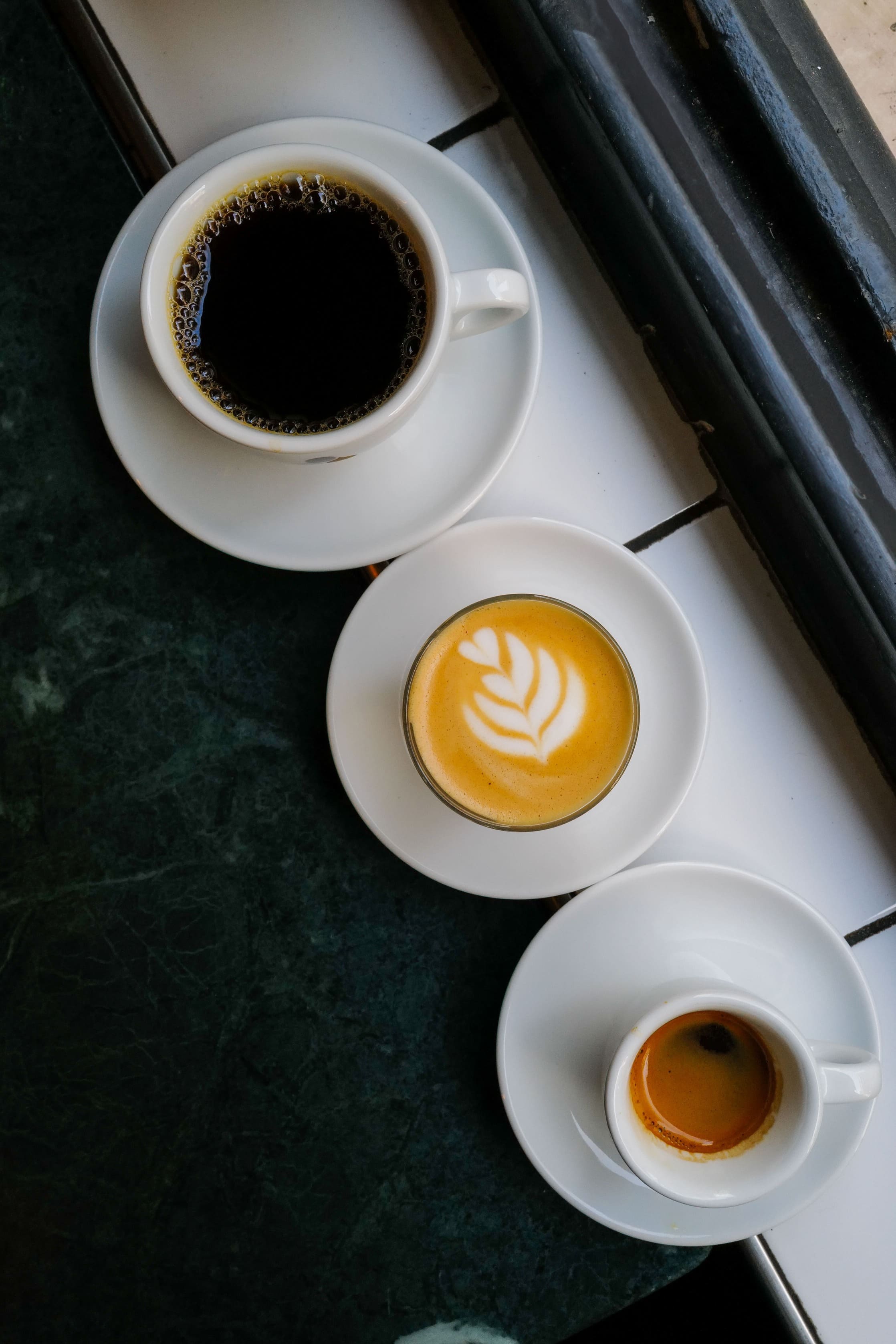Vue de dessus de trois tasses de café différentes — un café noir, un latte avec latte art et un espresso — alignées sur un rebord de fenêtre en carrelage blanc.